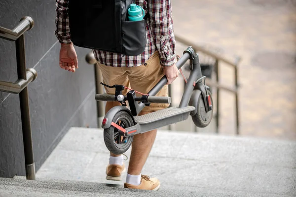 Katlanmış bir E-scooter taşıyan bir adam binanın yanındaki merdivenlerde. Çevresel şehir taşımacılığı, modern şehir konsepti. Ekolojik teknolojik yaşam tarzı. Katlanmış Konumda Elektrikli Scooter Taşıyıcı.