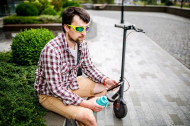 Elektronik scooter kullanan ve şehirde akıllı telefon kullanan bir adam. Elektro scooterlı modern bir erkek cep telefonuyla sohbet ederken parkta bankta oturuyor. Eko Taşımacılık öğrencisi..