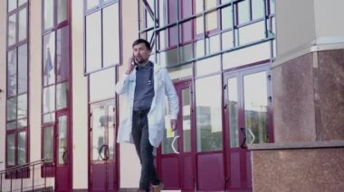 Steteskoplu profesyonel tıp doktoru hastaneden çıkarken cep telefonuyla konuşuyor. Gözlüklü ve beyaz cüppeli genç bir doktor, klinikten çıkarken telefonla konuşuyor.