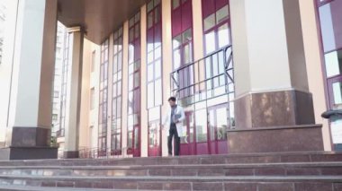 Steteskoplu profesyonel tıp doktoru hastaneden çıkarken cep telefonuyla konuşuyor. Gözlüklü ve beyaz cüppeli genç bir doktor, klinikten çıkarken telefonla konuşuyor.