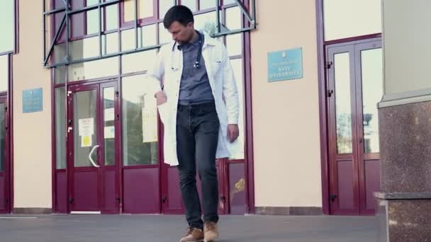 Молодой доктор в халате говорит по мобильному телефону, покидает здание больницы по лестнице. Медицинский работник покидает больницу по мобильному телефону. Студенты медицинских вузов выходят на улицу с помощью смартфона — стоковое видео