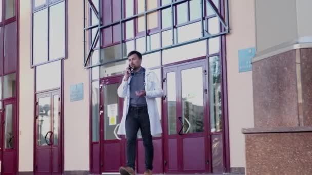 Медицинская профессиональная тема. Молодой доктор в белом халате покидает здание больницы, разговаривая по мобильному телефону с пациентом. Студенты медицинского университета пользуются смартфоном — стоковое видео