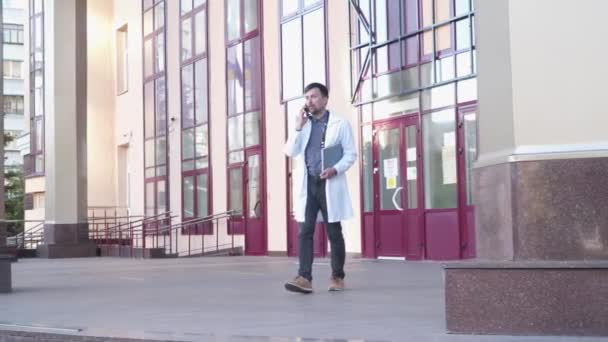 Medizin Gesundheitsberuf Thema. Junger Arzt im weißen Laborkittel verlässt das Krankenhausgebäude, während er mit dem Patienten telefoniert. Medizinstudentin vor Eingang nutzt Smartphone — Stockvideo