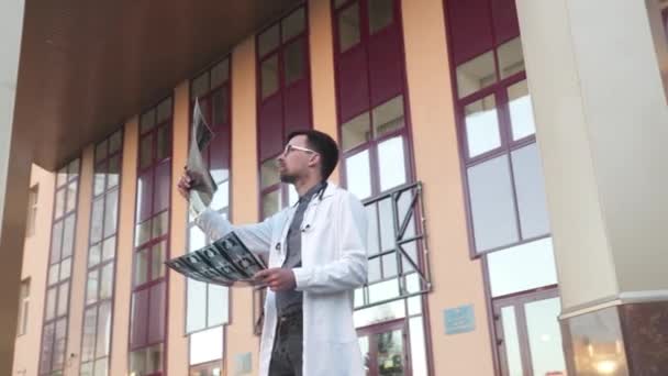 Młody lekarz, stażysta w białym fartuchu laboratoryjnym, stoi przy głównym wejściu do szpitala z prześwietleniem lub tomografią komputerową. Radiolog badający tomografię na zewnątrz. Zapalenie płuc, koncepcja wirusa korony — Wideo stockowe