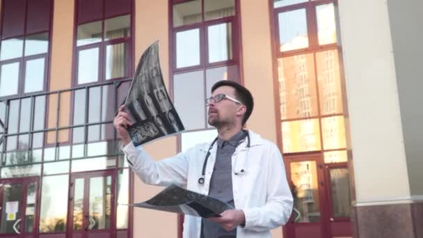 Medicina e saúde tema. Estudante de medicina examina uma tomografia computadorizada de pulmões fora da entrada da universidade. Jovem médico examinando radiografias de um paciente coronavírus. Médico analisa a ressonância magnética — Vídeo de Stock