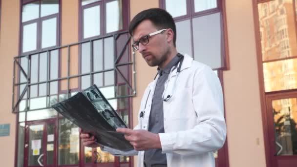 Een jonge arts, een stagiaire in een witte labjas, staat bij de hoofdingang van het ziekenhuis met een röntgen- of CT-scan. Radioloog onderzoekt MRI scan buiten. Longontsteking, corona virus concept — Stockvideo