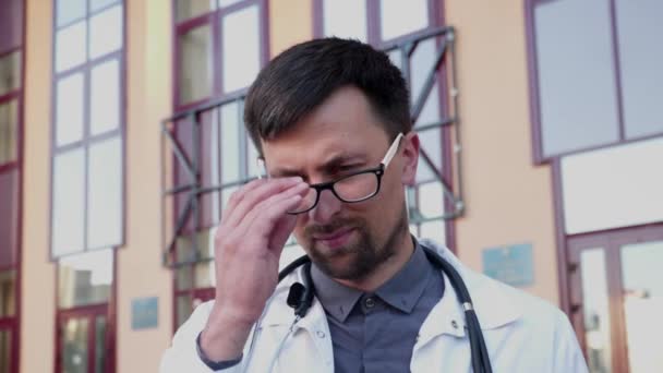 Overworked doctor takes off his glasses while leaving clinic for break near main entrance of building. Emaciated male doctor holding glasses, touching eyes and having headache outside the hospital — Stock Video