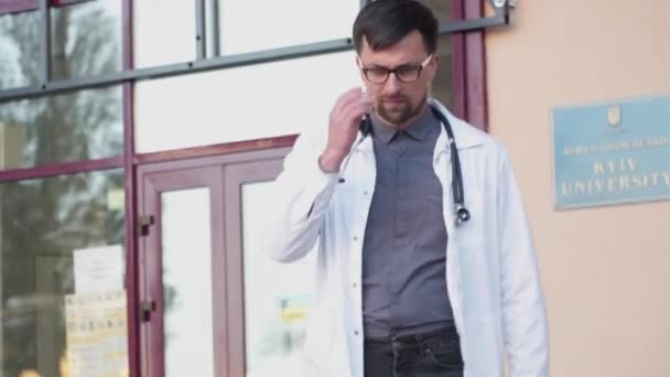 Overworked doctor takes off his glasses while leaving clinic for break near main entrance of building. Emaciated male doctor holding glasses, touching eyes and having headache outside the hospital — Stock Video