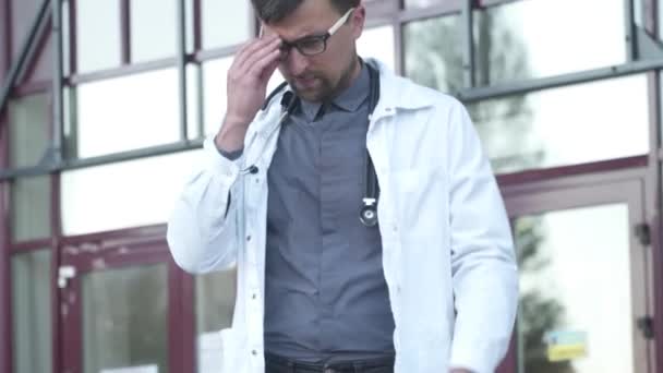 Overworked doctor takes off his glasses while leaving clinic for break near main entrance of building. Emaciated male doctor holding glasses, touching eyes and having headache outside the hospital — Stock Video