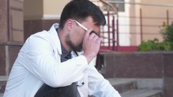 Overworked doctor takes off his glasses while leaving clinic for break near main entrance of building. Emaciated male doctor holding glasses, touching eyes and having headache outside the hospital — Stock Video