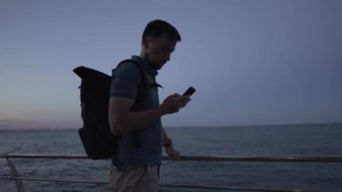 Adam akşam karanlığında modern akıllı telefon kamerasıyla denizin doğal ortamının fotoğraflarını çekiyor. Sırt çantalı gezgin cep telefonu kullanıyor. Uygulamayla deniz suyu yapıyor.