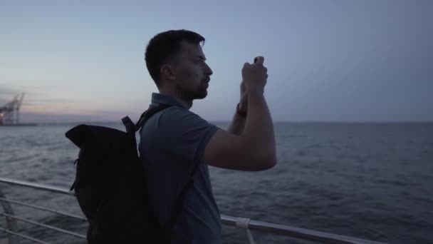 Homem tira fotos no crepúsculo da noite do ambiente natural do mar usando a câmera de smartphone moderna, viajante cara com mochila segurando telefone celular, fazendo foto água do mar através de um aplicativo — Vídeo de Stock