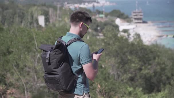 Man checking his cell phone during hiking trip. Young male using his mobile phone gps while out hiking along the path along the sea on a hill. Traveler view route on navigator in smart phone app map — Stock Video