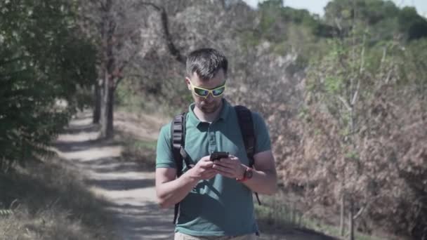 Caucasian man walks along trail on hill along sea, one-day hiker uses the Internet and smartphone app to navigate. Male builds a route in phone using satellites gps for orientation and geolocation — Stock Video