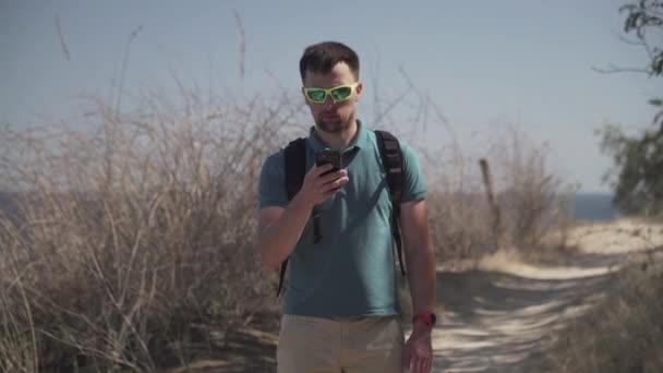 人は海の近くの山の歩道でGPS装置を使用して昼間のハイキング中にスマートフォンを使用します。男性ハイカーは海岸沿いのパス上の携帯電話上の地図アプリで正しいルートをチェックします — ストック動画