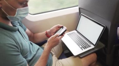 Telefondaki adam dizüstü bilgisayarla mesaj atıyor. Koronavirüs salgını sırasında banliyö treninde maske takıyor. Tema trenle seyahat, COVID 19 sırasında yeni kurallar. Erkek trende akıllı telefon ve dizüstü bilgisayar kullanır