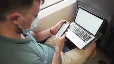 Trende çalışan, hava kirliliğini, tozu, PM2.5 pm2 'yi, koronavirüsü veya COVID 19' u önlemek için maske takan bir adam. Akıllı telefonda internette sörf yapan ve pencerenin yanında dizüstü bilgisayar kullanan bir erkek. Demiryolu taşımacılığı