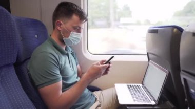 Telefondaki adam dizüstü bilgisayarla mesaj atıyor. Koronavirüs salgını sırasında banliyö treninde maske takıyor. Tema trenle seyahat, COVID 19 sırasında yeni kurallar. Erkek trende akıllı telefon ve dizüstü bilgisayar kullanır