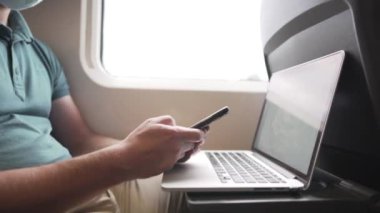 Telefondaki adam dizüstü bilgisayarla mesaj atıyor. Koronavirüs salgını sırasında banliyö treninde maske takıyor. Tema trenle seyahat, COVID 19 sırasında yeni kurallar. Erkek trende akıllı telefon ve dizüstü bilgisayar kullanır