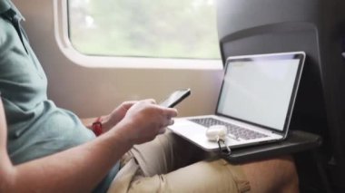 Trende çalışan, hava kirliliğini, tozu, PM2.5 pm2 'yi, koronavirüsü veya COVID 19' u önlemek için maske takan bir adam. Akıllı telefonda internette sörf yapan ve pencerenin yanında dizüstü bilgisayar kullanan bir erkek. Demiryolu taşımacılığı