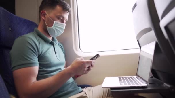 Transporte ferroviário e transporte de passageiros covid 19 pandemia. Homem caucasiano em máscara médica protetora em seu rosto segura o telefone na mão e usa laptop enquanto sentado junto à janela do trem durante a viagem — Vídeo de Stock