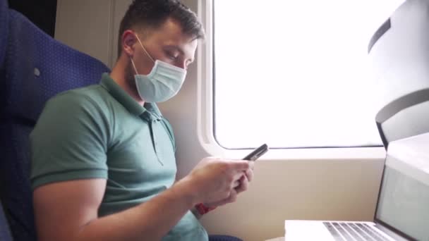 主题是旅行时的工作。年轻的白人男性自由职业者坐在靠窗的火车车厢里，一边用智能手机发短信，一边带着防护口罩在笔记本电脑上工作 — 图库视频影像