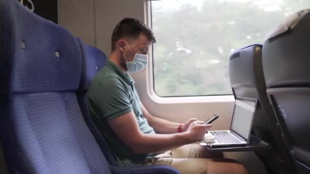 Мужчина пишет смс по телефону с ноутбуком, пригородный поезд в защитной маске во время пандемии коронавируса. Путешествие по железной дороге, новые правила жизни во время COVID 19. Мужчина использует смартфон и ноутбук в поезде — стоковое видео