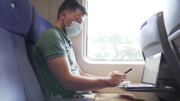 電車の中で働く男は、空気汚染、ほこり、 PM2.5 pm2 、コロナウイルスやCOVID 19を防ぐためにマスクを着用しています。スマートフォンでインターネットをサーフィンし、ウィンドウの近くにラップトップを使用して男性の乗客。鉄道輸送 — ストック動画