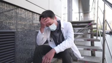 Endişeli ve yorgun doktor hastane merdivenlerinde oturuyor. Beyaz önlüklü ve tıbbi koruyucu maskeli bitkin bir doktor merdiven kliniğinde oturuyor. COVID-19 sırasındaki stres sağlık çalışanı