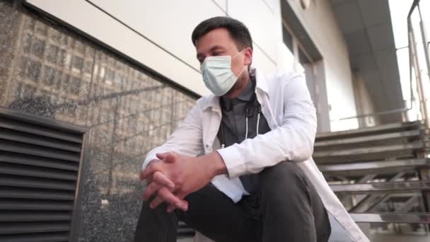Кавказский молодой врач садится на лестницу возле здания клиники, усталый и несчастный, потирая нос и глаза, чувствуя усталость и головную боль. Концепция стресса и разочарования работников здравоохранения — стоковое видео