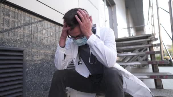 Ernsthafter Arzt ruht nach langer Schicht im Krankenhaus auf der Treppe. Müder Arzt in Maske, weißer Uniform und Stethoskop, während er auf der Treppe vor der Klinik sitzt, Kopfschmerzen und sich die Augen reibt — Stockvideo