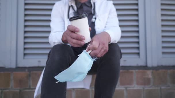 Primo piano di mano con tazza di caffè e maschera medica protettiva. Stanco, medico esausto in camice bianco medico durante la pausa caffè all'aperto ospedale al mattino. COVID-19, pandemia di coronavirus — Video Stock