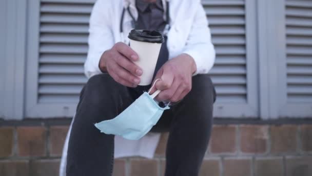 Medicina, salud y concepto pandémico. Triste joven médico agotado, mano de cerca con taza de papel de café desechable para llevar y máscara médica, sentado fuera del patio trasero del hospital — Vídeos de Stock