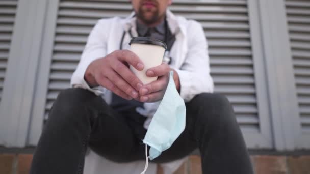커피 한잔과 보호용 의료용 마스크로 손을 클로즈업 했습니다. 아침에 야외 병원에서 휴식 시간에 커피를 마시다가 지친 채로 하얀 가운을 입고 있는 의사. 코로나 바이러스 대유행 COVID-19 — 비디오