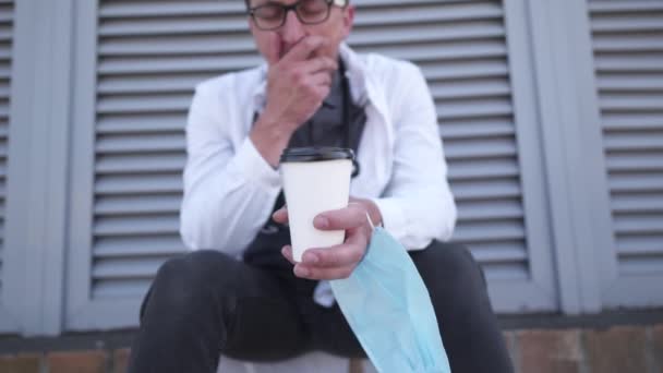 Tıp, sağlık ve salgın konsepti. Üzgün, yorgun, genç bir erkek doktor, tek kullanımlık kahve fincanı ve tıbbi maskeyle hastanenin arka bahçesinde oturuyor. — Stok video