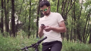 Dağ bisikleti süren spor giyimli bir erkek bisiklet bilgisayarında gezinmek için bir cihaz kullanıyor. Ormanda spor yapan bir adam, tahta üzerinde bisiklete biniyor ve elinde GPS navigatörü tutuyor. Guy uygulama ekran haritasına dokun
