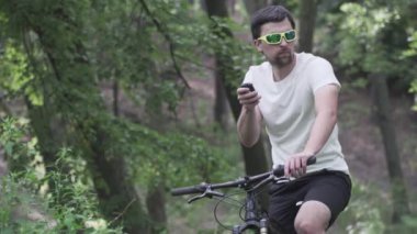 Dağ bisikleti süren spor giyimli bir erkek bisiklet bilgisayarında gezinmek için bir cihaz kullanıyor. Ormanda spor yapan bir adam, tahta üzerinde bisiklete biniyor ve elinde GPS navigatörü tutuyor. Guy uygulama ekran haritasına dokun