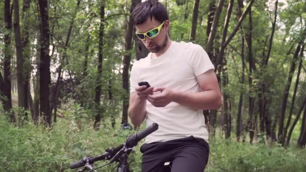 Dağ bisikleti süren spor giyimli bir erkek bisiklet bilgisayarında gezinmek için bir cihaz kullanıyor. Ormanda spor yapan bir adam, tahta üzerinde bisiklete biniyor ve elinde GPS navigatörü tutuyor. Guy uygulama ekran haritasına dokun — Stok video