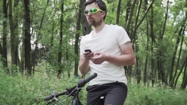 Muž ve sportovním oblečení na horském kole pomocí gadget navigovat na kole počítače. Muž atlet cvičení v lese, jízda na kole ve dřevě drží GPS navigátor v rukou. Mapa obrazovky Guy touch — Stock video