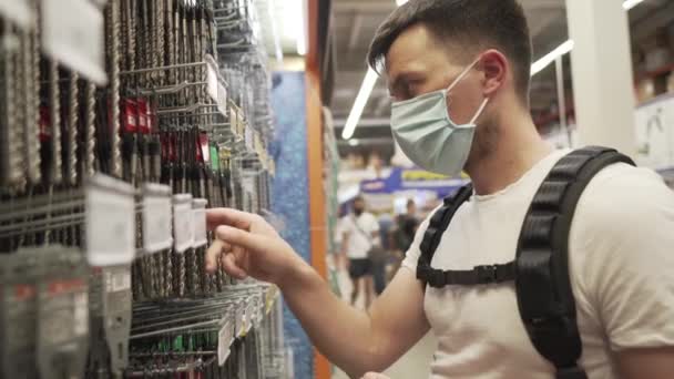 Кавказька людина в медичній масці вибирає вантаж буровий біт для будівельного обладнання в магазині. Мужній вибір металевого свердла встановлений для бурової машини в будівництві супермаркету протягом 19 пандемії — стокове відео