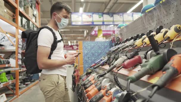 Mężczyzna kupujący wybierający elektryczne narzędzie ręczne w dziale supermarketu budowlanego w ochronnej masce medycznej. Pracownik warsztatu w sklepie z narzędziami kupuje narzędzie podczas covid 19 — Wideo stockowe