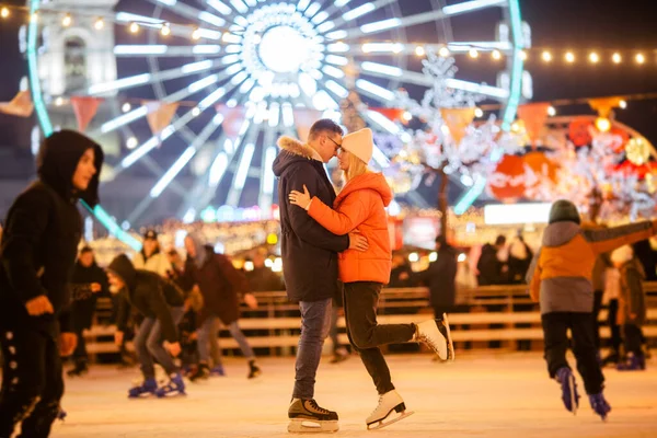 Man and woman in love in embrace on ice arena against of Ferris wheel in evening of Christmas. Couple at city skating rink, romance and glowing lanterns, light bulbs and garlands festive atmosphere.