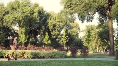 Park boyunca bisiklet yolunda elektrikli scooter süren genç bir adam. Şehir hayatı. Çevre taşımacılığı. Şehirdeki adam e-scooter 'a biniyor. Teknoloji, çevre dostu hareketlilik kavramı. Moda kentsel ulaşım