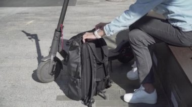 Kafkaslı yetişkin bir kadın şehirde bankta otururken elektrikli scooter kullandıktan sonra sırt çantasından güç bankasını alarak şarj ediyor. Dış güç bankasının pili şarj ediliyor