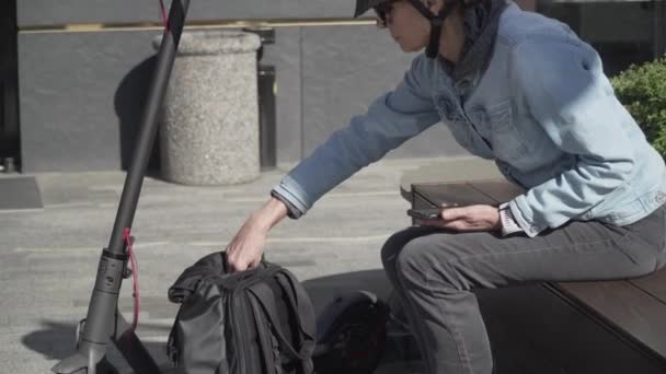 Dispositivos y dispositivos inteligentes modernos en la vida cotidiana persona urbana. Mujer adulta madura descansando en el banco junto a la scooter eléctrica y cargando el teléfono desde el cargador portátil powerbank tomándolo de la mochila — Vídeo de stock