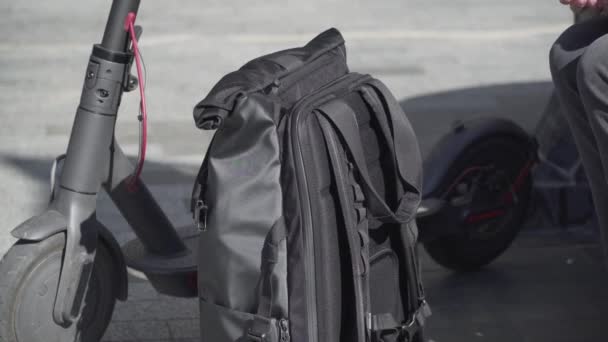 Взрослая белая женщина в шлеме берет банк питания из рюкзака, чтобы зарядить мобильный телефон после езды на электрическом скутере, сидя на скамейке в городе. Зарядка аккумулятора от внешнего блока питания — стоковое видео