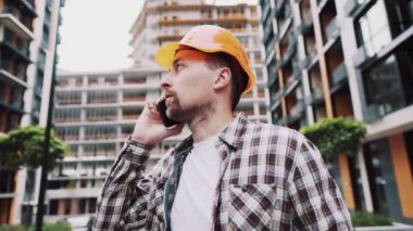 Turuncu kasketli ve ekose gömlekli beyaz erkek inşaat işçisi şantiyede telefonla konuşuyor. Mimarlık teması. Erkek işi. Foreman inşaat sürecini akıllı telefonla kontrol ediyor