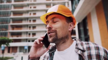 Turuncu kasketli ve ekose gömlekli beyaz erkek inşaat işçisi şantiyede telefonla konuşuyor. Mimarlık teması. Erkek işi. Foreman inşaat sürecini akıllı telefonla kontrol ediyor