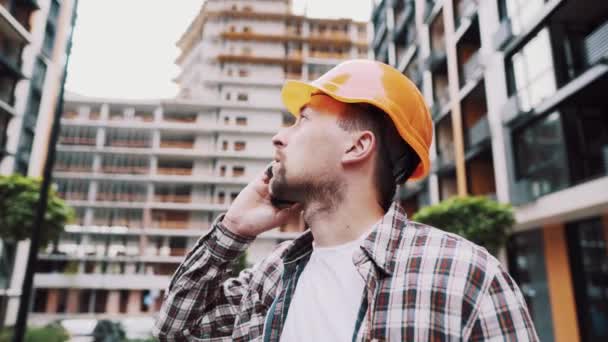 Portrait of young handyman making call while standing at construction area. Engineer talking on the phone on a construction site. Builder in helmet control according to plan by mobile phone — Stock Video