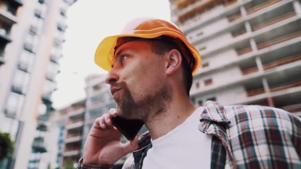 Portret van een jonge klusjesman die belt terwijl hij in de bouw staat. Ingenieur aan de telefoon op een bouwplaats. Bouwer in helm controle volgens plan door mobiele telefoon — Stockvideo